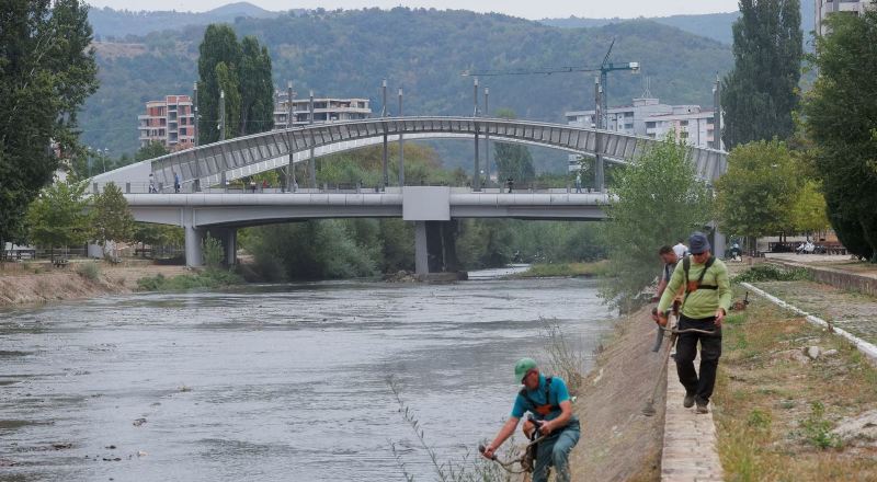 Punëtorë duke kositur bar buzë lumit Ibër, afër urës kryesore të qytetit të ndarë etnikisht, në Mitrovicë, më 9 shtator 2024. Reuters.